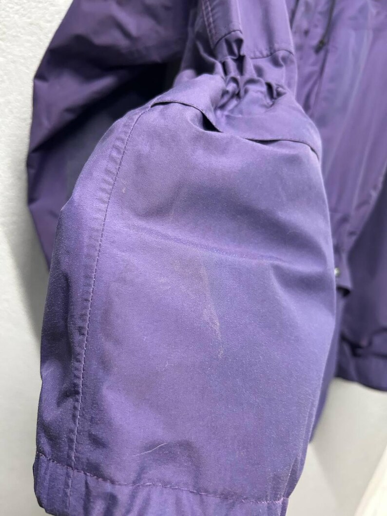 May include: Close-up of a purple jacket sleeve. The sleeve is gathered at the cuff and has a smooth, slightly shiny texture. The stitching is visible along the seams. The jacket appears to be made of a lightweight, water-resistant material.