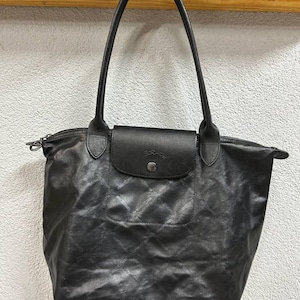以下が含まれることがあります： 繰り返し模様のあるテクスチャー仕上げの黒いトートバッグ。中央にはスナップボタン付きのフラップがあり、2本の細いショルダーストラップが付いています。バッグの上部にはファスナーがあり、構造化された台形のシルエットをしています。