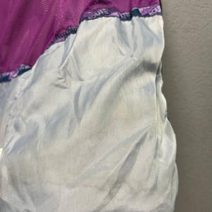 May include: Close-up of a purple and gray jacket interior. The jacket features a mesh lining in a vibrant purple color, contrasted by a light gray fabric. The bottom hem is a deep navy blue, with a drawstring and a black toggle.