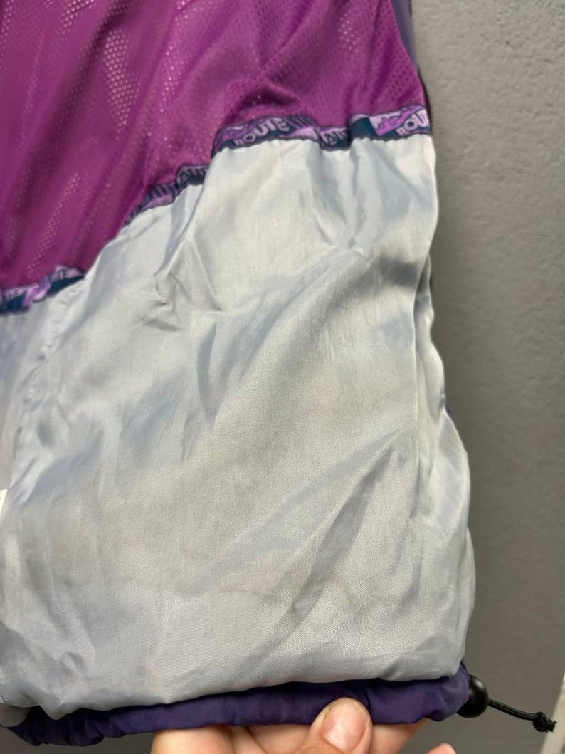 May include: Close-up of a purple and gray jacket interior. The jacket features a mesh lining in a vibrant purple color, contrasted by a light gray fabric. The bottom hem is a deep navy blue, with a drawstring and a black toggle.