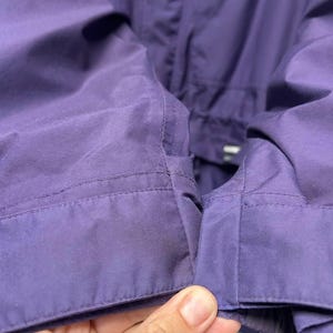May include: Close-up of a section of a purple jacket. The fabric is smooth and appears to be a lightweight material. The stitching is visible along the seams, and the jacket has a tailored appearance. The color is a deep, saturated purple.