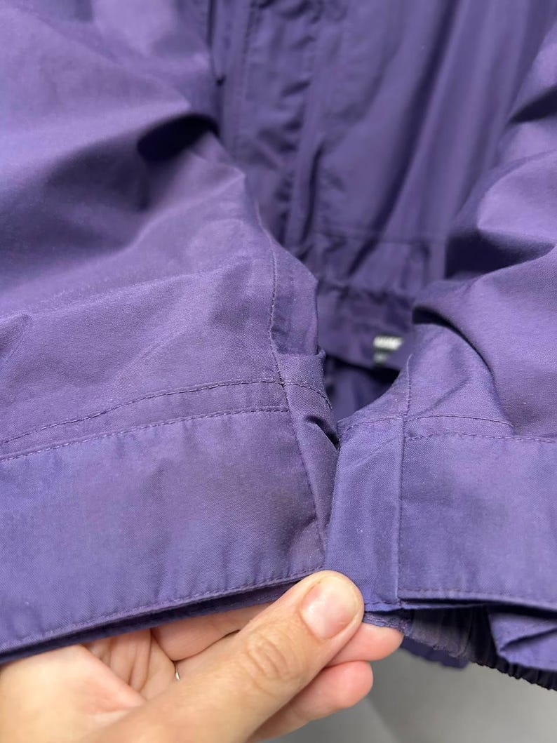 May include: Close-up of a section of a purple jacket. The fabric is smooth and appears to be a lightweight material. The stitching is visible along the seams, and the jacket has a tailored appearance. The color is a deep, saturated purple.