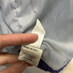 May include: A close-up of a tag inside a garment. The tag reads "MADE IN ITALY" and lists the material as 100% Poliestere, Polyester, and Polyester. The garment's interior is light blue.