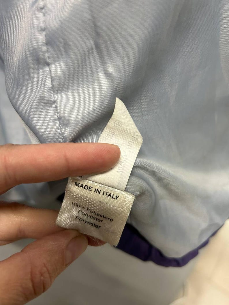 May include: A close-up of a tag inside a garment. The tag reads "MADE IN ITALY" and lists the material as 100% Poliestere, Polyester, and Polyester. The garment's interior is light blue.