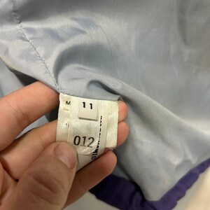 May include: Close-up of a garment's interior, showing a label with the number "11" and the number "012". The garment's lining is a light blue color, with a purple trim. The label is white with black text.