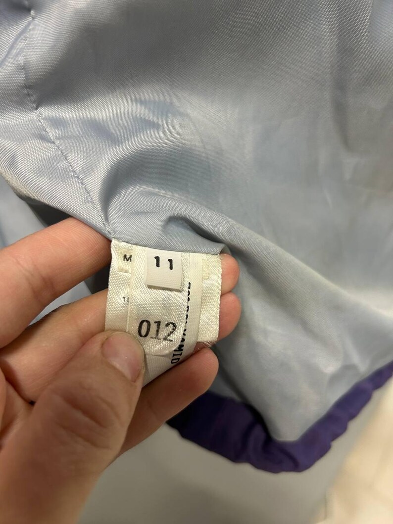 May include: Close-up of a garment's interior, showing a label with the number "11" and the number "012". The garment's lining is a light blue color, with a purple trim. The label is white with black text.