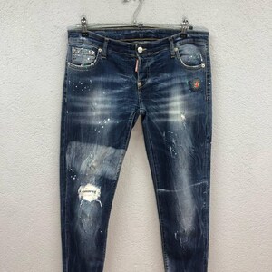 May include: Dark wash denim jeans with a distressed look, featuring paint splatter details and a ripped knee. The jeans are hanging on a silver hanger against a neutral background. The jeans have a button closure and pockets.