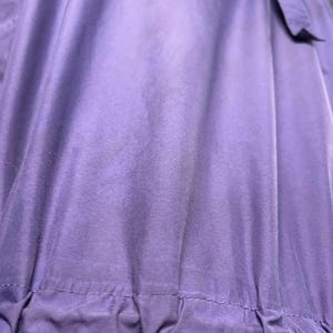 May include: Close-up of a purple garment with a gathered waistline and a dark purple accent. The fabric has a sheen, and the garment appears to be made of a lightweight material. A bow is visible at the top.
