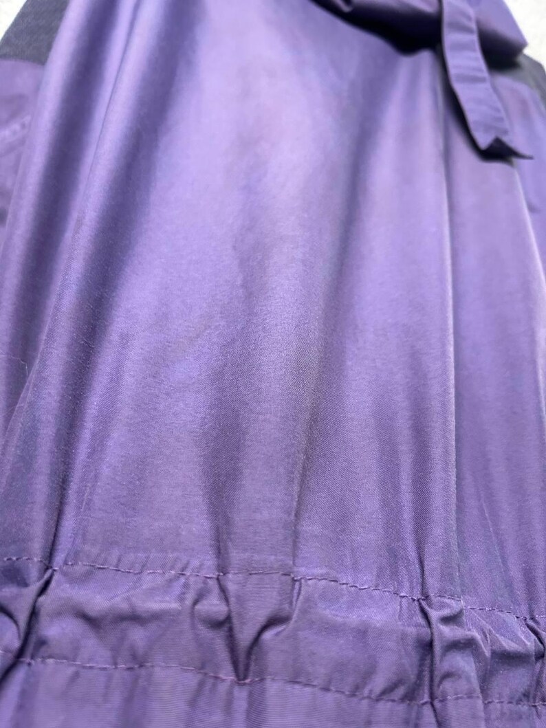 May include: Close-up of a purple garment with a gathered waistline and a dark purple accent. The fabric has a sheen, and the garment appears to be made of a lightweight material. A bow is visible at the top.