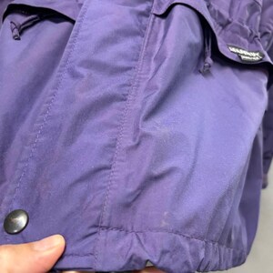 May include: Close-up of a purple jacket with multiple pockets and a drawstring hem. The jacket has a black button and a label with white text. The fabric appears to be water-resistant. The jacket is a solid color.