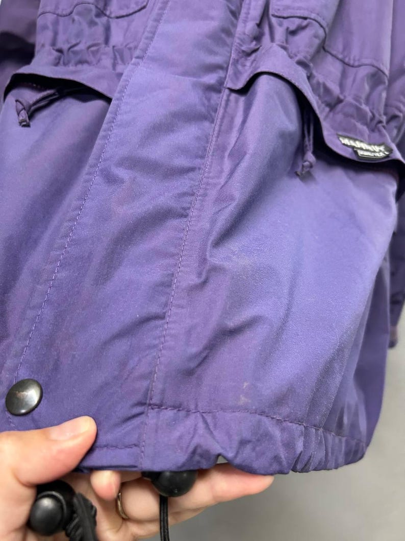 May include: Close-up of a purple jacket with multiple pockets and a drawstring hem. The jacket has a black button and a label with white text. The fabric appears to be water-resistant. The jacket is a solid color.