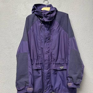 May include: A purple and gray hooded jacket with a drawstring waist and multiple pockets. The jacket has gray shoulder and sleeve panels and the brand name "Mammut" is visible on the front. The jacket is hanging on a wooden hanger.