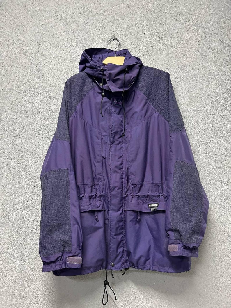 May include: A purple and gray hooded jacket with a drawstring waist and multiple pockets. The jacket has gray shoulder and sleeve panels and the brand name "Mammut" is visible on the front. The jacket is hanging on a wooden hanger.