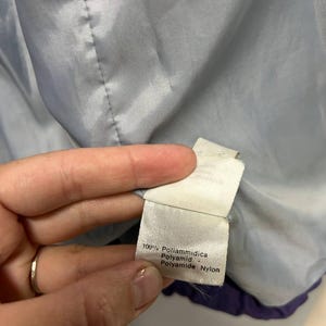 May include: Close-up of a white tag with text in Italian and English, including "100% Poliammidica" and "Polyamide Nylon." The tag is attached to a light blue and purple garment lining.