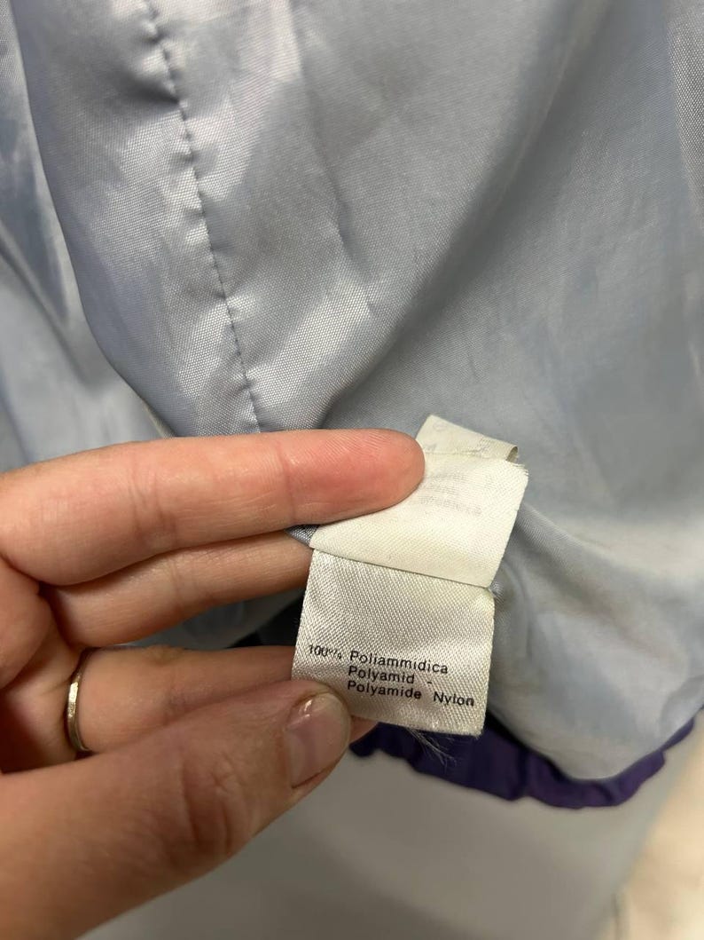May include: Close-up of a white tag with text in Italian and English, including "100% Poliammidica" and "Polyamide Nylon." The tag is attached to a light blue and purple garment lining.