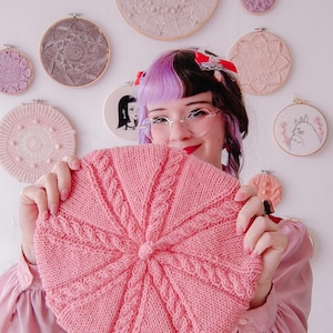 Puede incluir: Un boina rosa tejida con un diseño de punto trenzado. La boina se sostiene frente a una persona. Aros de bordado con varios diseños son visibles en el fondo.