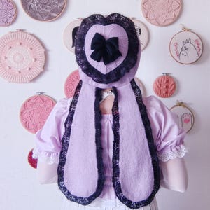 May include: A lilac-colored knitted hood with a heart-shaped design and a black bow. The hood is trimmed with black lace and has long, scarf-like extensions. The background features various embroidered hoops in shades of pink and purple.