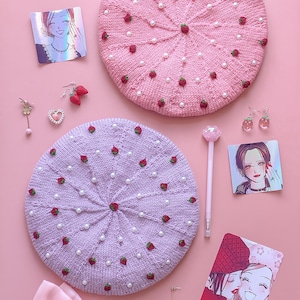 Puede incluir: Dos boinas de punto rosa y morado con adornos de perlas y fresas. Las boinas están rodeadas de lazos rosas, collares de perlas y otros accesorios.