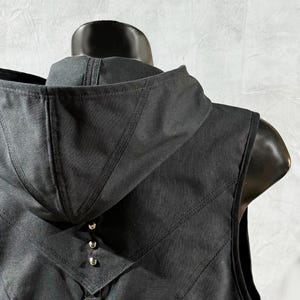 May include: Black sleeveless vest with a hood, featuring a black leather panel with a decorative chain and studs on the back.