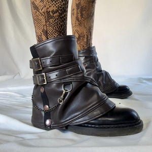 Pode incluir: Polainas de bota de couro marrom com fivelas e um detalhe de corrente. As polainas são usadas sobre botas pretas.