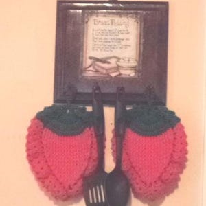 May include: Two crocheted strawberry-shaped pot holders in pink and green hanging on a black framed picture with a vintage recipe printed on it. The picture is hanging on a white wall. Two black kitchen utensils are hanging below the pot holders.