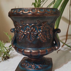 May include: A decorative urn with a square base, featuring a blue and bronze finish. The urn has handles and is adorned with grape vine relief details. Ideal for home decor, this piece adds a touch of classic elegance to any space.