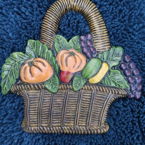 May include: A ceramic wall hanging in the shape of a basket overflowing with fruit. The basket is brown with a woven texture. The fruit includes oranges, a red apple, a yellow pear, and grapes.