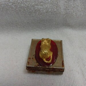 May include: A small, square, metal trinket box with a red oval on the top. The oval has a gold-coloured cat figurine on it.