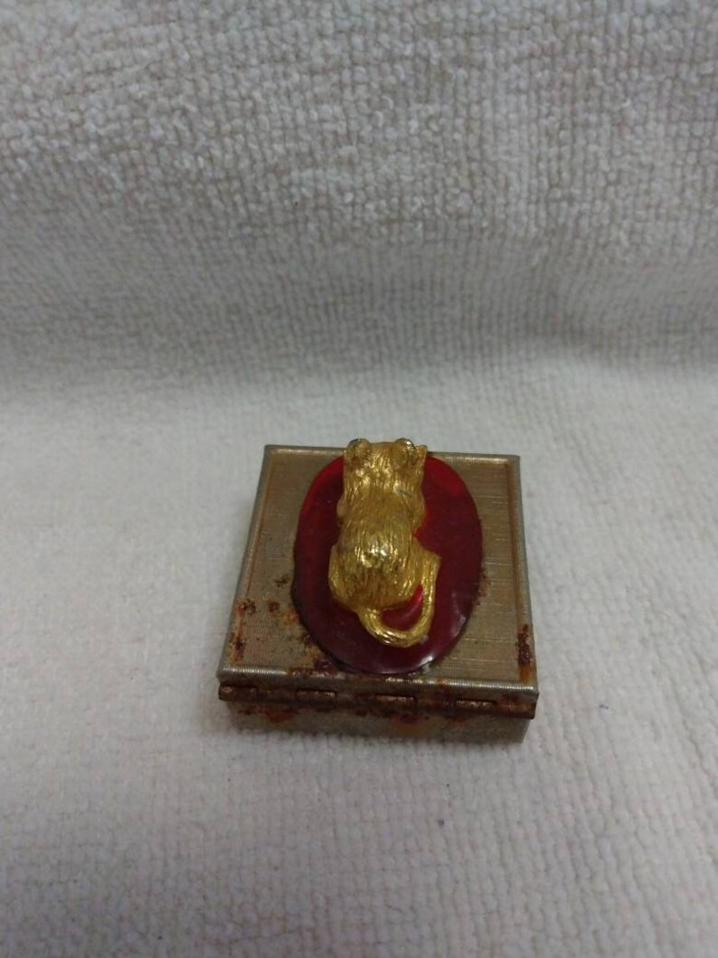 May include: A small, square, metal trinket box with a red oval on the top. The oval has a gold-coloured cat figurine on it.