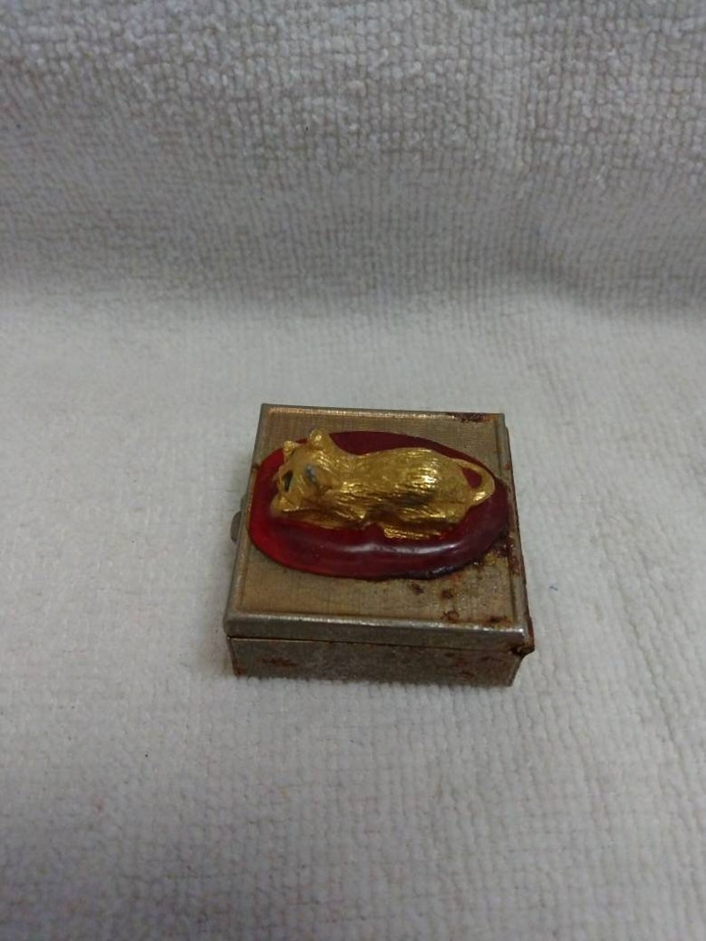 May include: A small, rectangular, metal box with a red interior and a gold-coloured cat figurine on the lid.