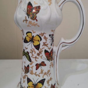 May include: White ceramic pitcher with a tall, slender design. The pitcher is adorned with colorful butterfly illustrations and gold accents. The handle and rim are also trimmed with gold. A decorative piece for the home.