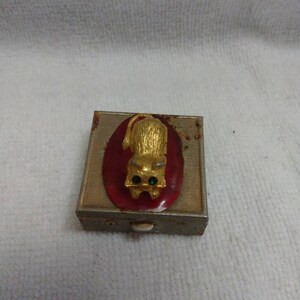 May include: A small, rectangular, metal trinket box with a red oval on the front. The oval features a gold cat with green eyes. The box has a hinged lid and a small, white button on the front.