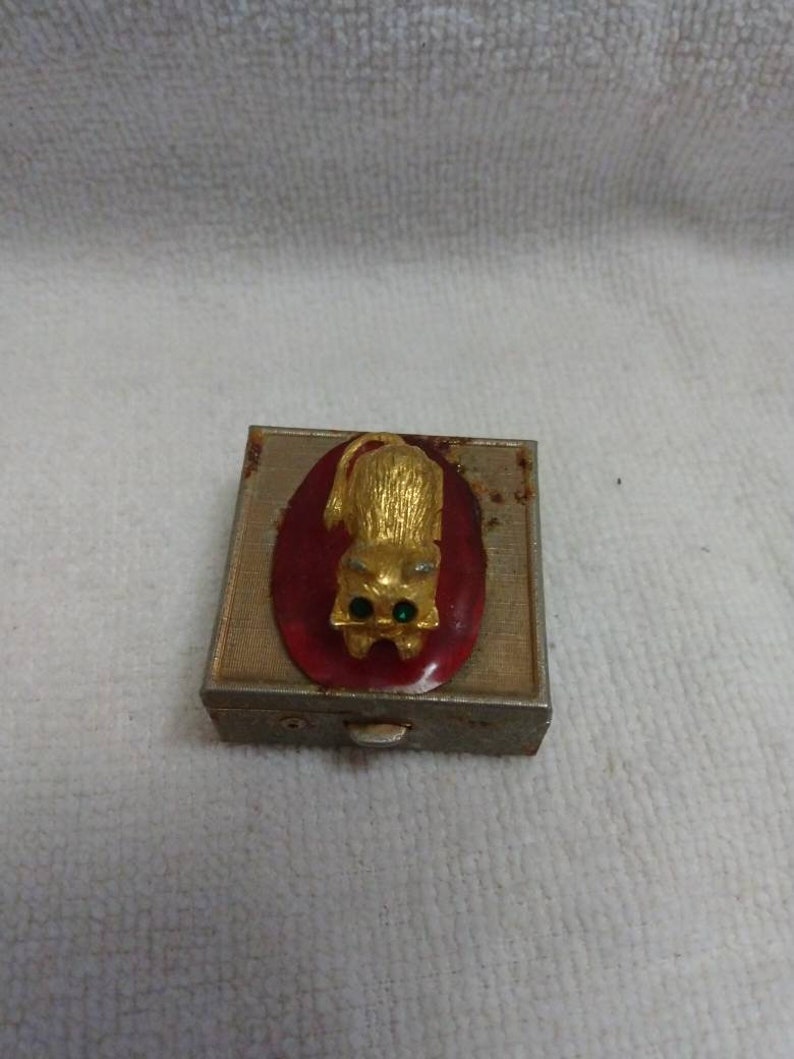 May include: A small, rectangular, metal trinket box with a red oval on the front. The oval features a gold cat with green eyes. The box has a hinged lid and a small, white button on the front.