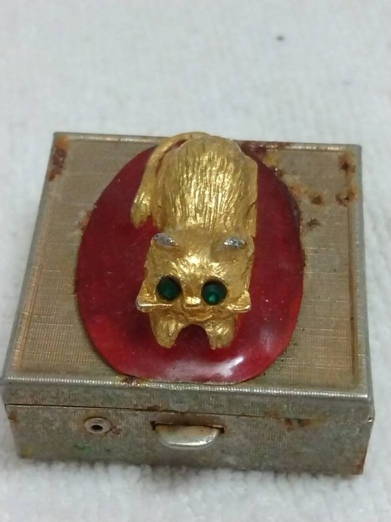 May include: A vintage metal trinket box with a gold cat figurine on the lid. The cat has green gem eyes and is sitting on a red oval.