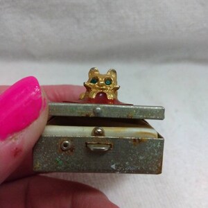 May include: A small, vintage metal trinket box with a hinged lid. The box is decorated with a gold-coloured cat figurine with green gem eyes. The box has a worn, aged appearance.