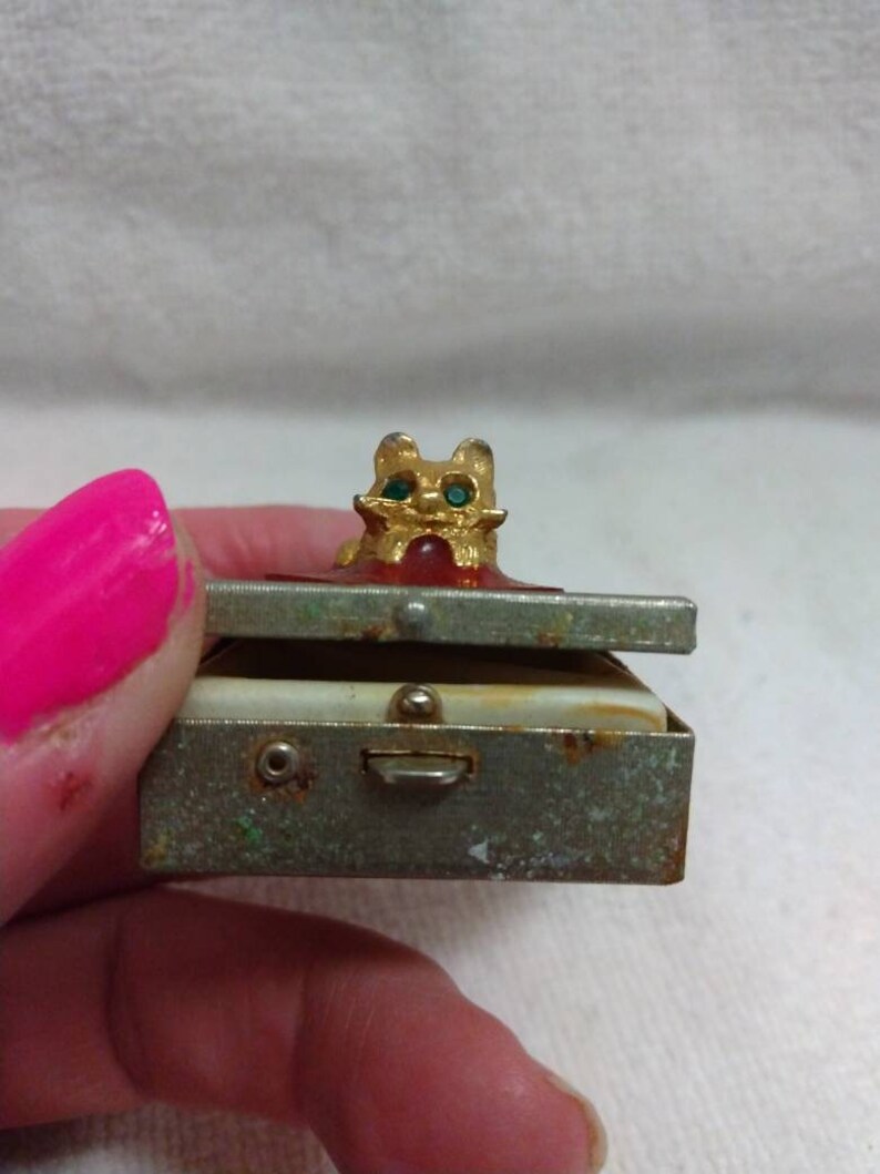 May include: A small, vintage metal trinket box with a hinged lid. The box is decorated with a gold-coloured cat figurine with green gem eyes. The box has a worn, aged appearance.