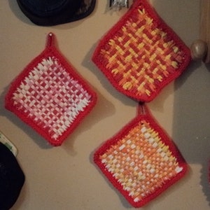 May include: Three red and white crocheted pot holders with a woven pattern. The pot holders are hanging on a wall.