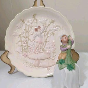 May include: A decorative ceramic plate and bell featuring fairy-themed designs. The plate has a raised relief of a fairy on a mushroom among flowers. The bell is white with a fairy figure holding a green stem. The plate is supported by a gold stand.