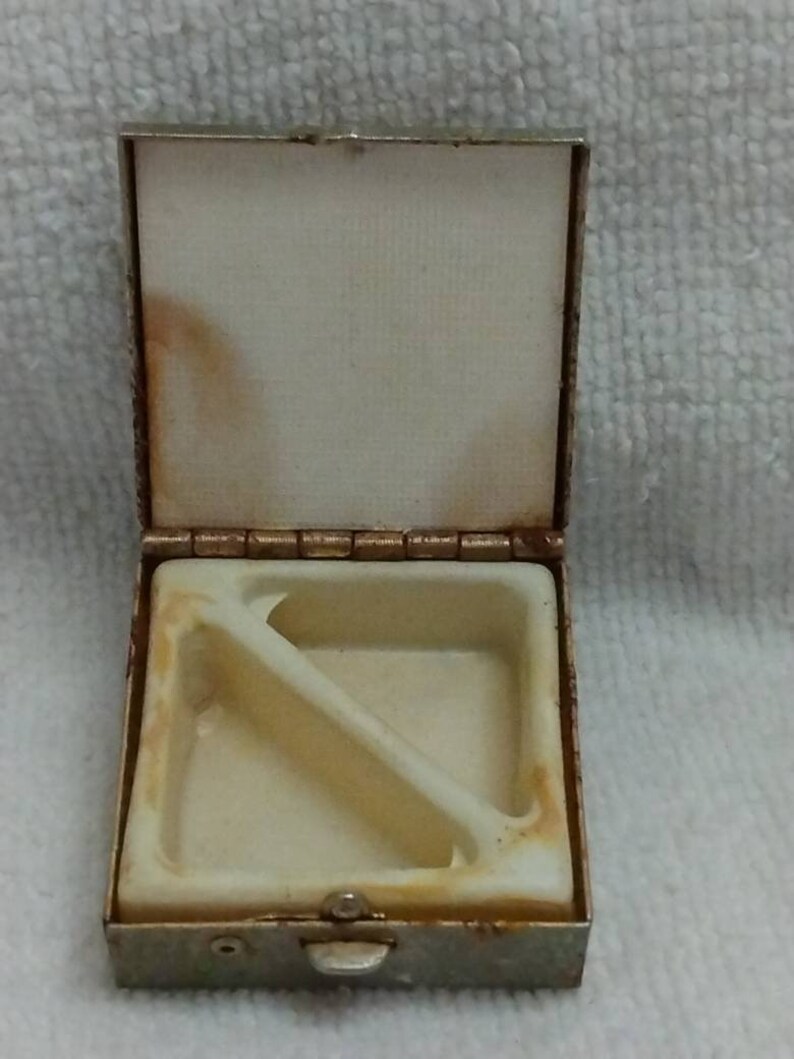 May include: A small, silver-coloured metal box with a hinged lid. The box is divided into two compartments by a diagonal divider. The box is slightly tarnished and has a worn appearance.