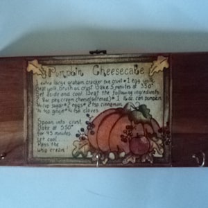 May include: A wooden box with a recipe for pumpkin cheesecake printed on the front. The recipe includes ingredients like graham cracker crust, cream cheese, pumpkin, eggs, sugar, cinnamon, and cloves. The box has two hooks on each side.