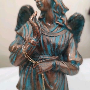 May include: A bronze-colored angel statue with turquoise accents. The figure is kneeling, with outstretched hands and detailed wings. A gold-colored chain hangs from the statue's hands. The statue has a distressed finish.