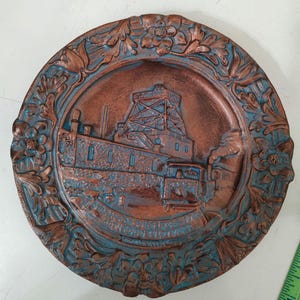 May include: A copper-colored metal plate with a blue-green patina. The plate features a raised image of a building with a tower and a train. The plate is surrounded by a decorative border of flowers and leaves.