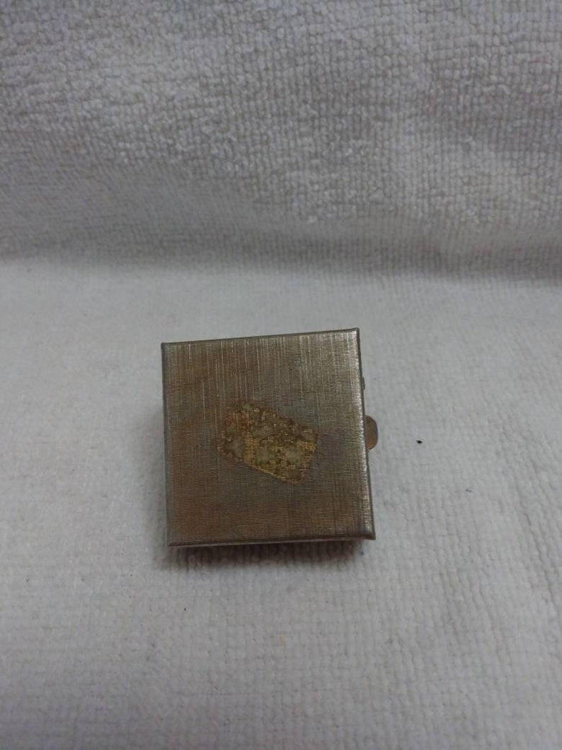 May include: A small, silver, rectangular box with a gold design on the top. The box has a hinged lid and a small latch.