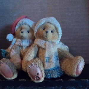 May include: Two plush teddy bears wearing scarves and hats. The bear on the left is wearing a red and white Santa hat and a white scarf. The bear on the right is wearing a blue and white scarf and a white hat.