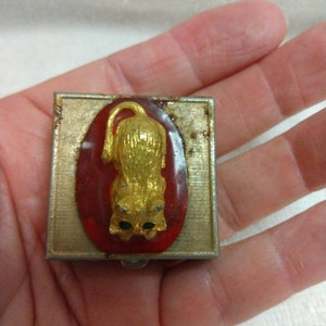 May include: A small, square, silver-coloured metal box with a red and gold cat design. The cat is sitting on a red oval with gold accents. The box has a hinged lid.