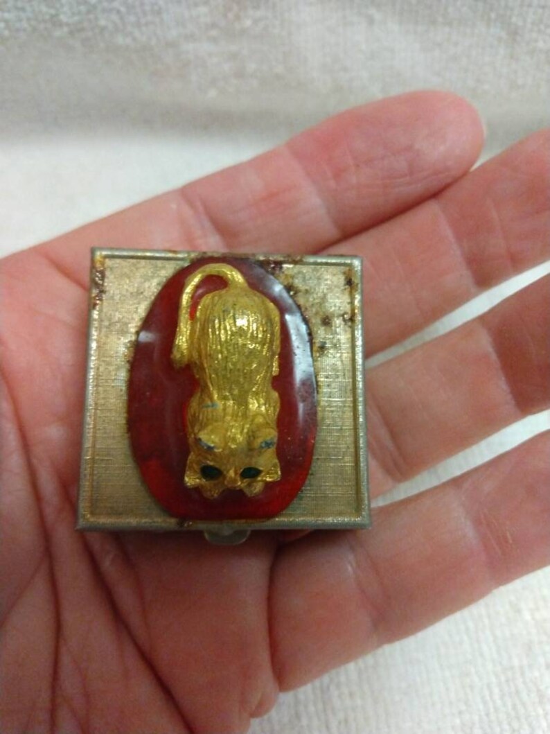 May include: A small, square, silver-coloured metal box with a red and gold cat design. The cat is sitting on a red oval with gold accents. The box has a hinged lid.