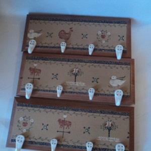 May include: Three wooden wall-mounted key racks with a brown background and a decorative floral and animal design. Each rack has five white ceramic hooks.