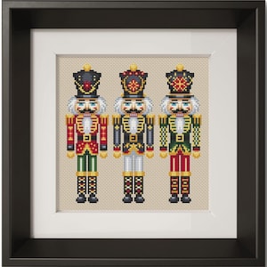 May include: A framed cross-stitch embroidery featuring three nutcracker figures standing side by side. Each nutcracker wears a tall hat, a detailed uniform with gold accents, and white hair. The figures are rendered in red, green, blue, and gold.