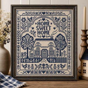 May include: A framed cross-stitch sampler featuring a dark blue house design on a light beige fabric background. The text HOME SWEET HOME is centered above the house, surrounded by decorative borders of birds, trees, flowers, and small hearts.