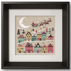 Puede incluir: Obra de arte en punto de cruz enmarcada que representa una escena de un pueblo invernal. Papá Noel en su trineo tirado por renos vuela sobre casas coloridas, una luna creciente y copos de nieve. La obra está enmarcada en un marco negro con un paspartú blanco.